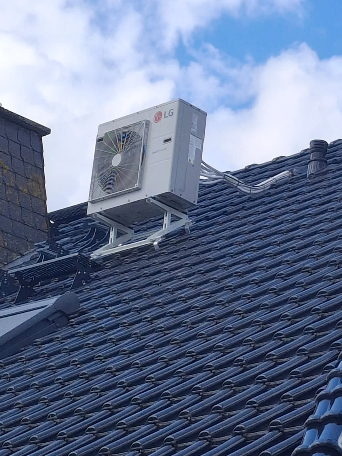 LG-Split-Klimaanlage: weißes Außengerät auf einer Metall-Dachhalterung über einem dunklen Ziegeldach vor blauem Himmel