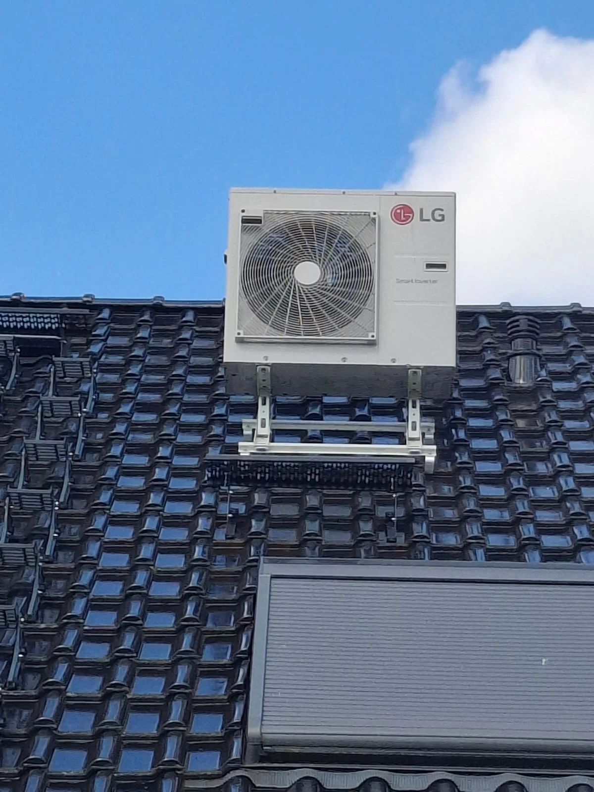 LG-Dual-Inverter-Außengerät weiß, frontal auf einer stabilen Metall-Dachhalterung über einem dunklen Ziegeldach vor blauem Himmel
