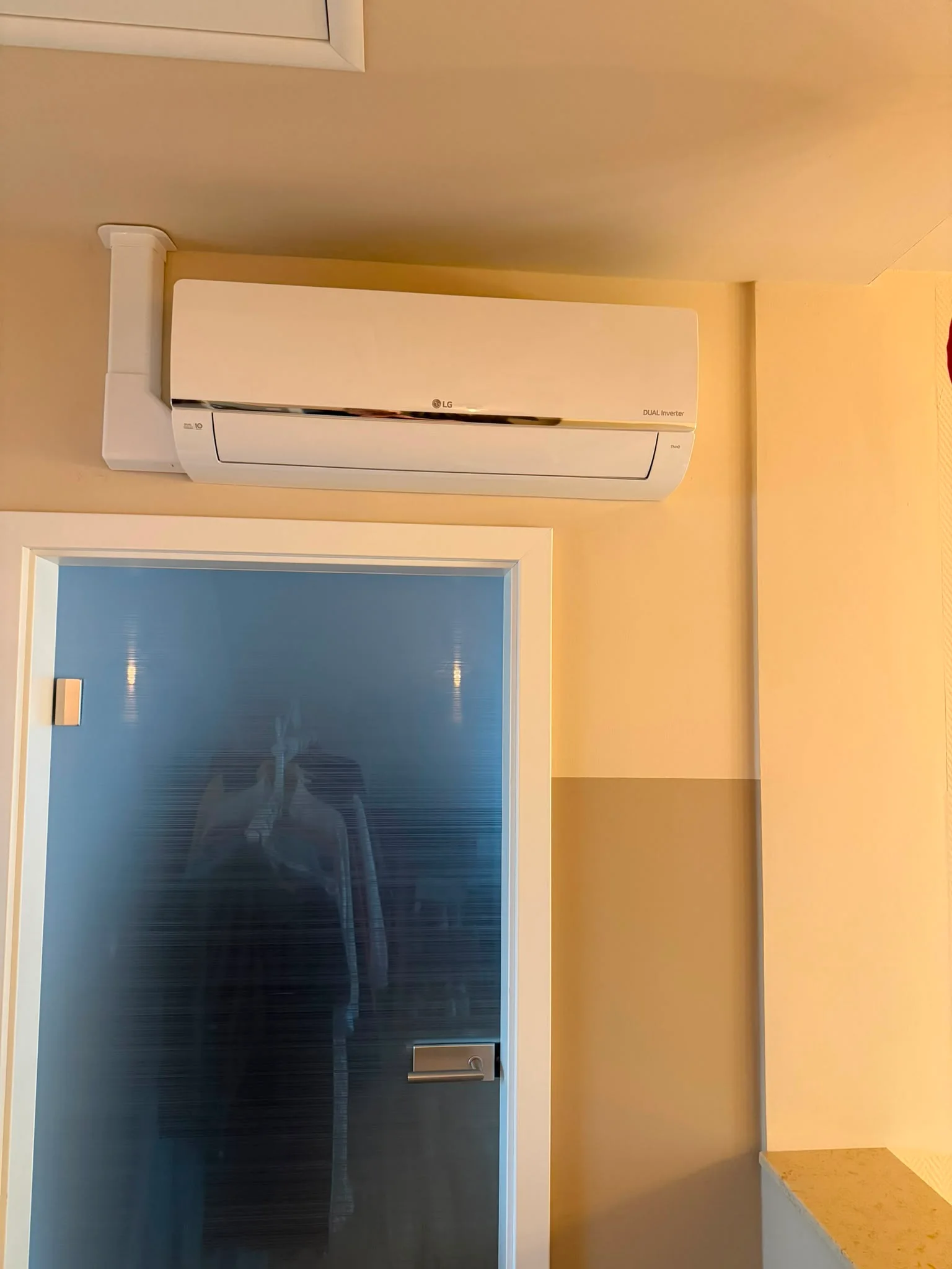 LG-Dual-Inverter-Innengerät weiß, wandmontiert oberhalb eines Türdurchgangs mit Glastür; die Leitungsführung verläuft in einem weißen Installationskanal senkrecht zur Decke