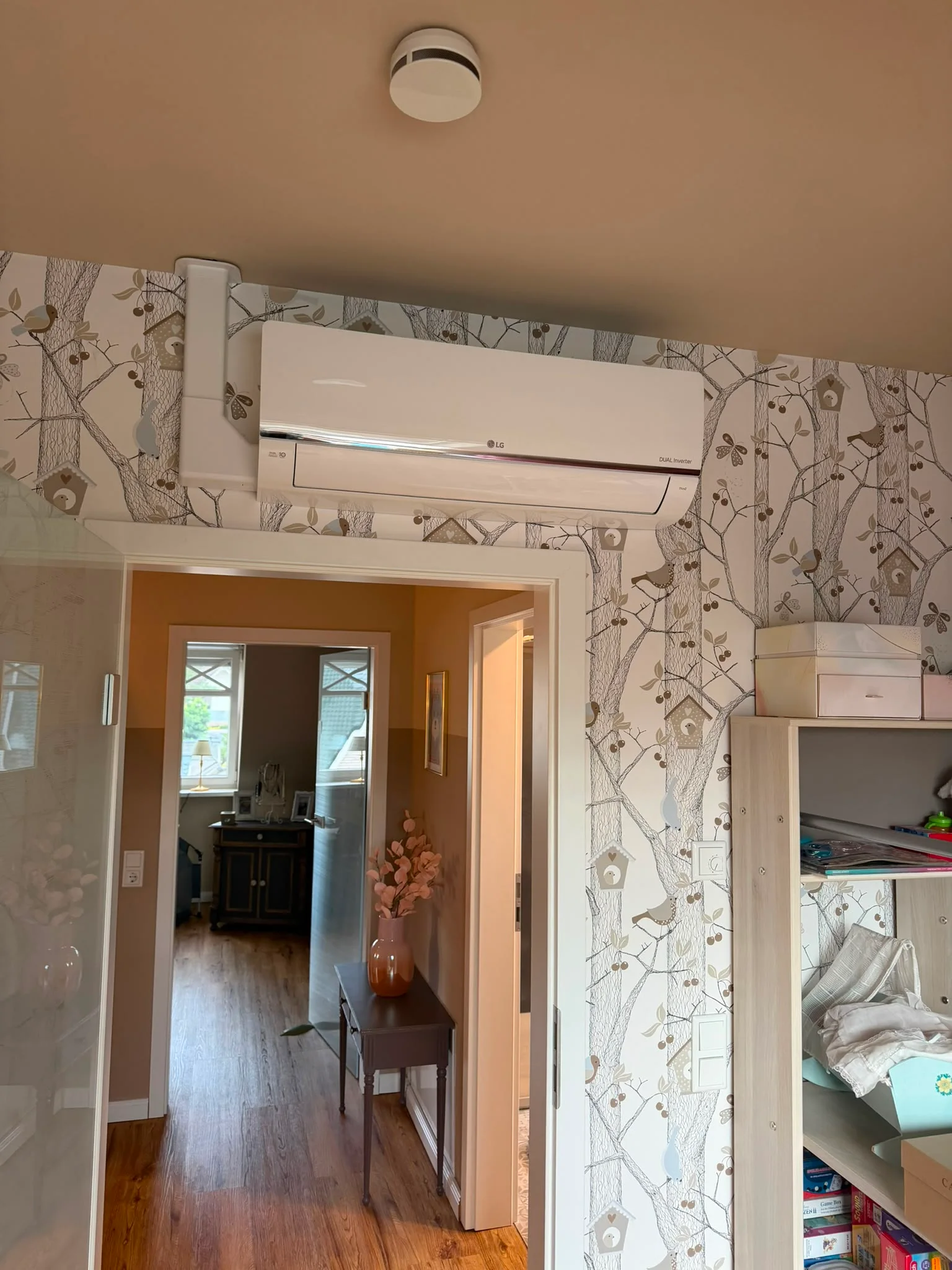 LG-Dual-Inverter-Innengerät weiß, montiert über einem Türdurchgang neben einer Tapete mit Baummotiv; weißer Installationskanal an der Wand