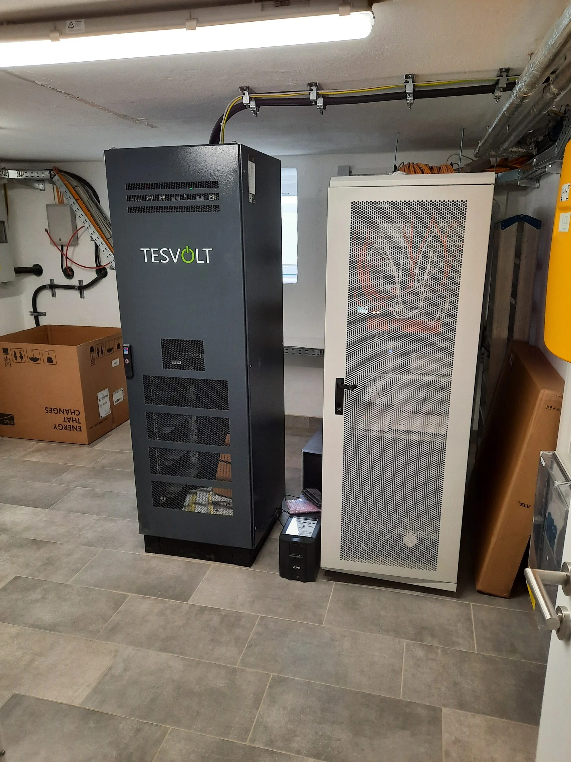 Dunkler Batteriespeicher-Schrank mit Belüftungsgitter neben einem hellen 19-Zoll-Serverschrank mit Lochblechtür im Technikraum, davor eine unterbrechungsfreie Stromversorgung auf gefliestem Boden