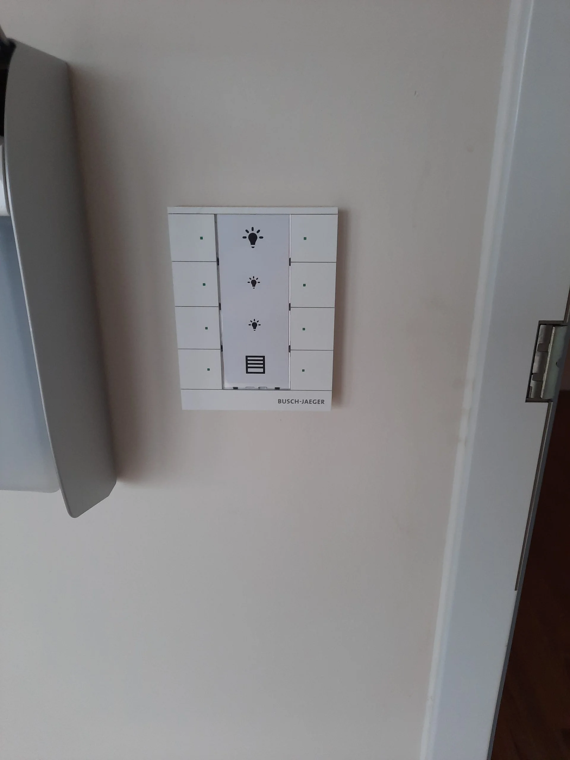 Weißes Busch-Jaeger Free@Home-Tastermodul an einer Wand mit Symbolen für drei Beleuchtungsszenen und Rollladensteuerung, daneben ein weißer Downlight-Deckenspot mit gekippter Blende