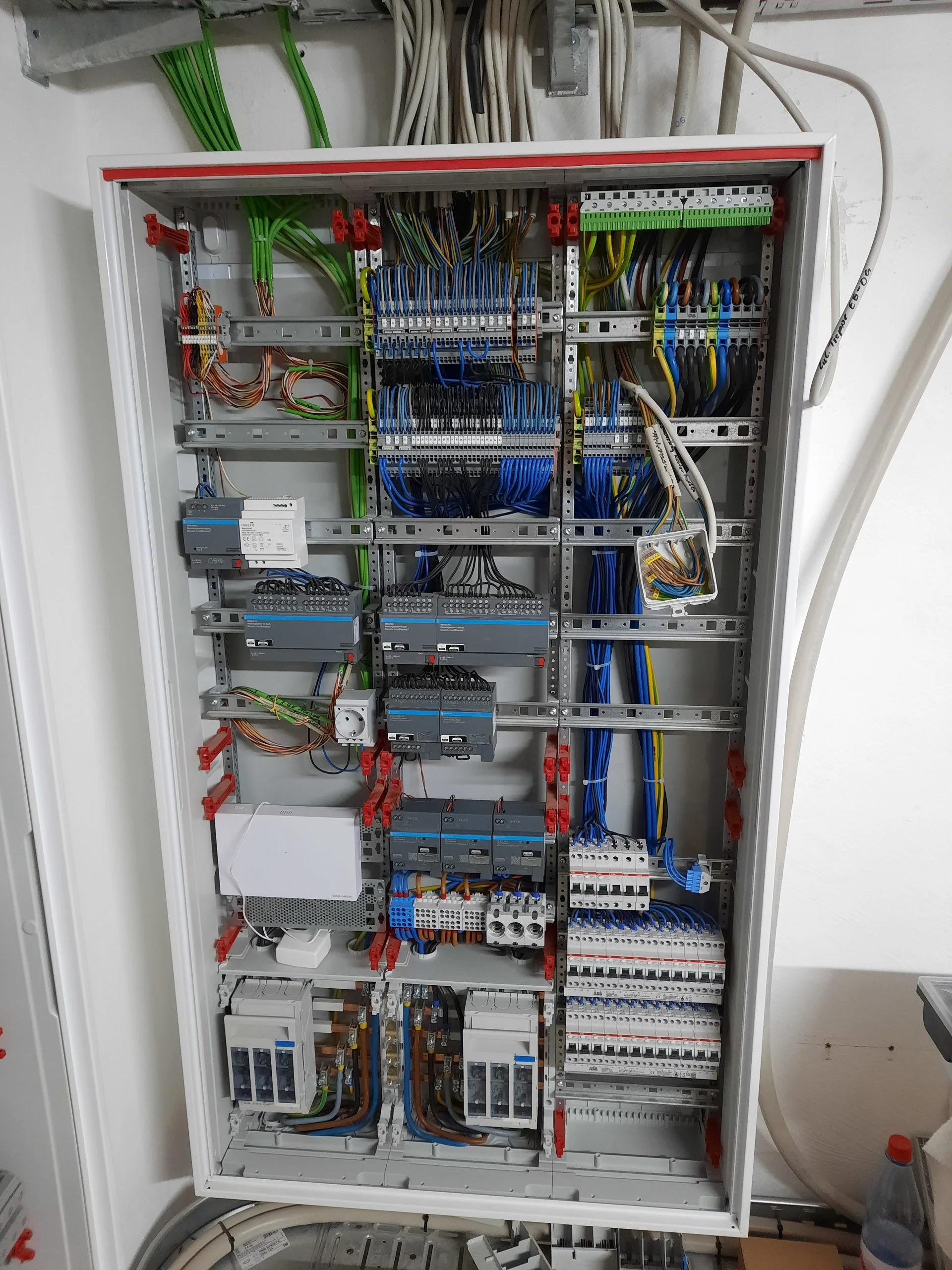 Geöffnete Elektro-Hauptverteilung im Technikraum mit mehreren Hutschienenreihen, Reihenklemmen, ABB-Leitungsschutzschaltern, Bus-Modulen und farblich sortierten Adern hinter blauen und grünen Zugentlastungen