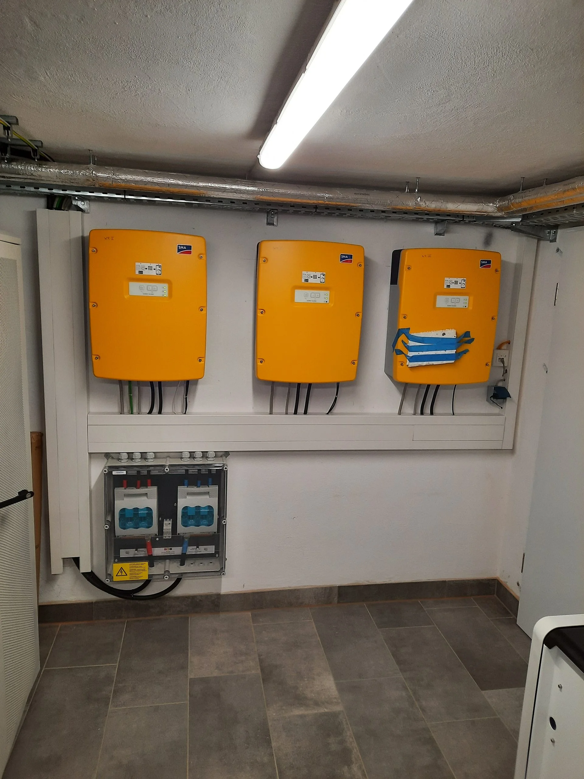 Drei orangefarbene SMA Sunny-Island-Batteriewechselrichter nebeneinander an der Wand des Technikraums, darunter ein transparenter DC-Anschlusskasten mit blauen Sicherungen und Zuleitungen