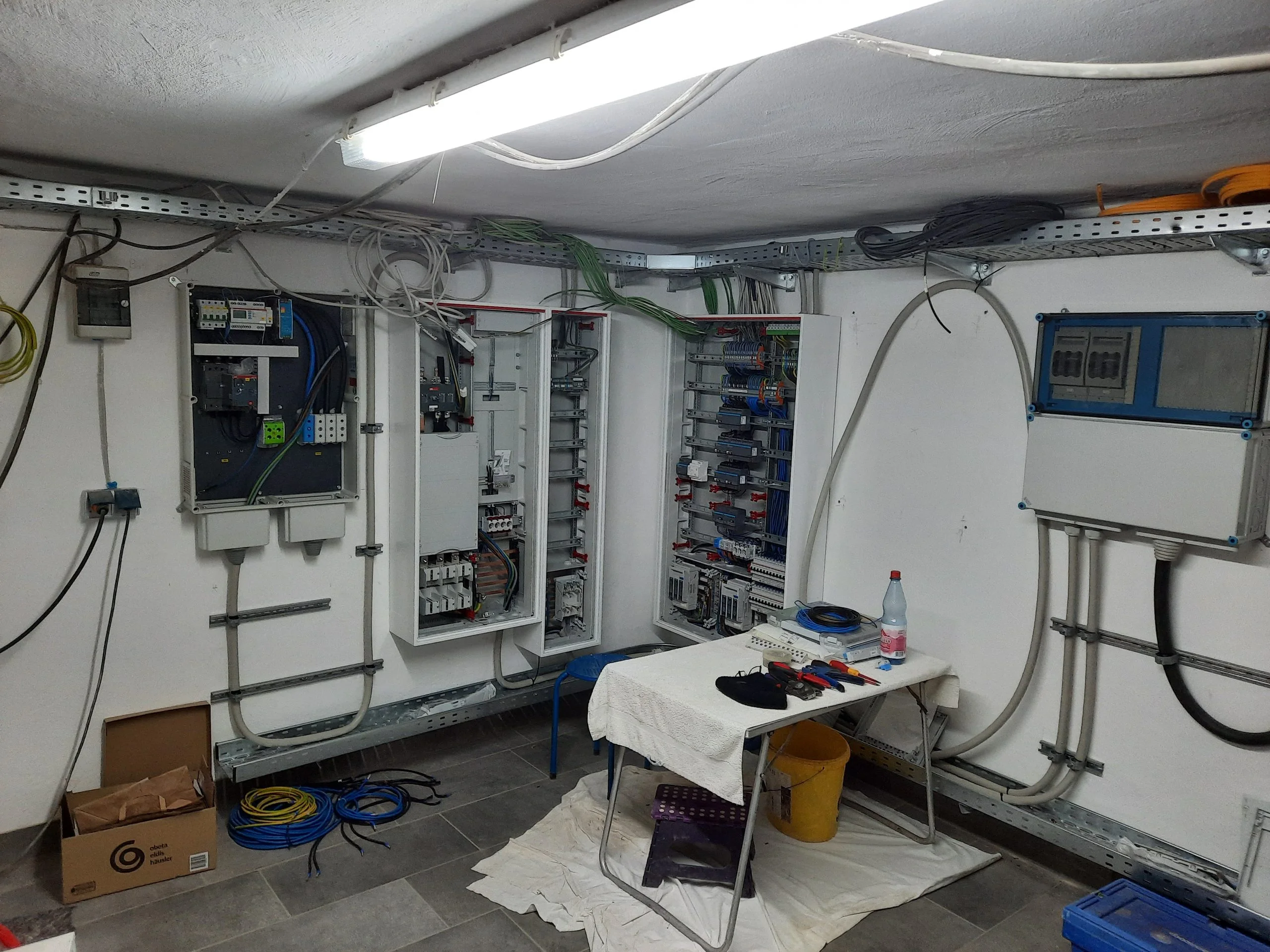 Technikraum eines Wohnobjekts in Trebitz bei Bad Belzig: mehrere geöffnete Elektroverteilungen mit Reihenklemmen und Schutzschaltern, PV-Wechselrichter, blauer Anschlusskasten, verzinkter Kabeltrassen-Ring an der Decke und Verlegematerial am Boden
